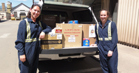 Two people in overalls standing by a truck with boxes and containers in the back.